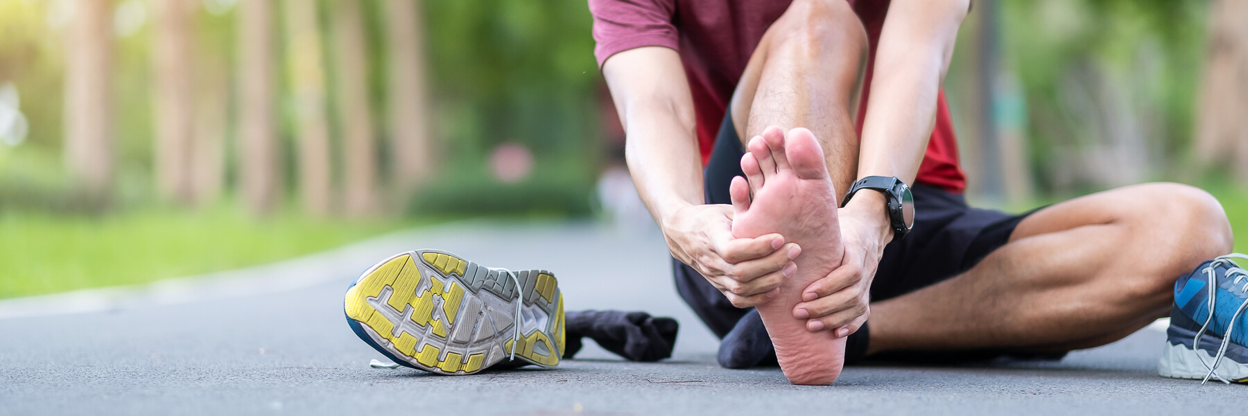 Young adult male sitting on ground with his running shoe off, holding foot or heel muscle pain, runner having heel or foot ache or pain due to Plantar fasciitis